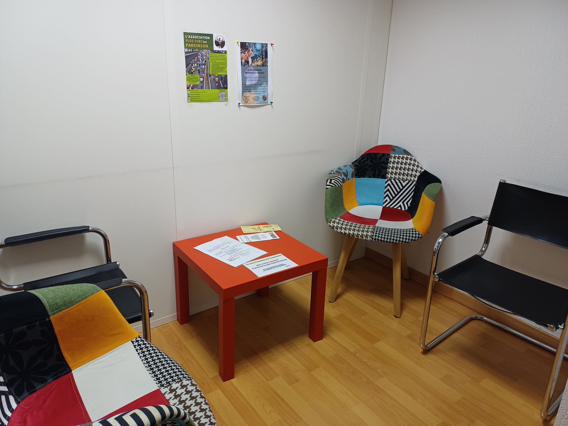 Petite salle d'attente avec des chaises dépareillées, une table rouge et des affiches sur le mur blanc.