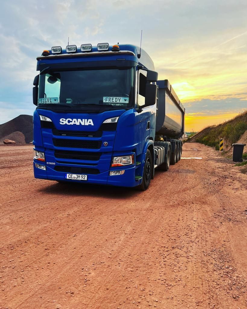 Blauer Scania-Sattelschlepper auf einer roten Schotterstraße, im Hintergrund ein Sonnenuntergang.
