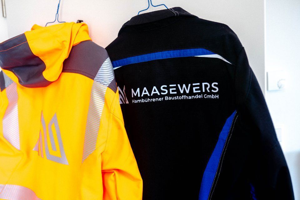 An einer Wand hängen eine gelbe Warnweste und eine schwarze Jacke mit dem Logo „Maasewers“.