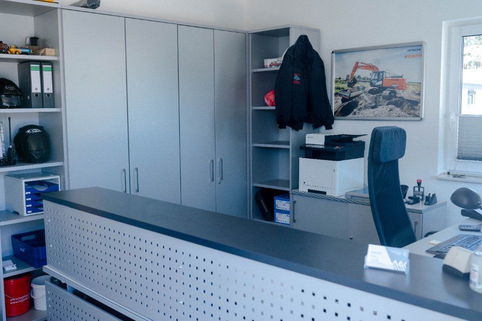 Büroeinrichtung: graue Schränke, Schreibtisch, Regale, Schreibtischstuhl, Jackenaufhängung, Bild von Baumaschinen.