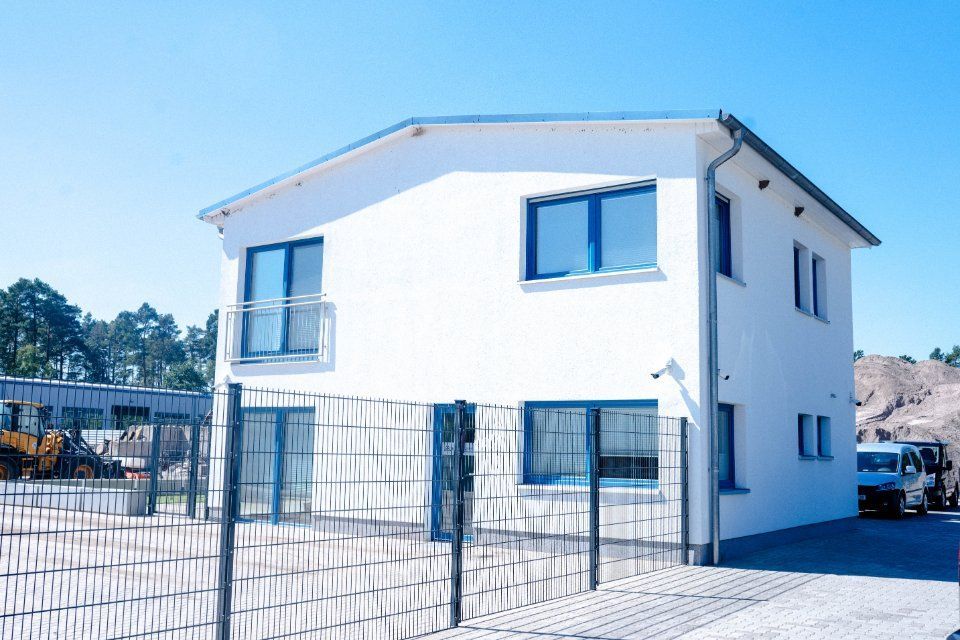 Weißes zweistöckiges Haus mit blau gerahmten Fenstern und einem kleinen Balkon vor einem Maschendrahtzaun.