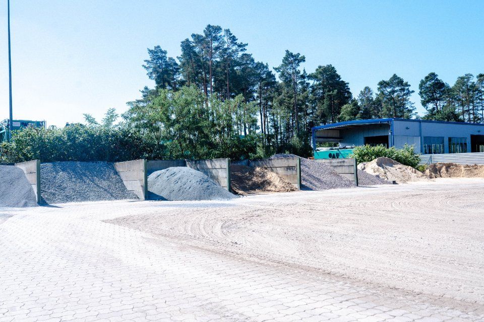 Mehrere Kieshaufen, möglicherweise auf einer Baustelle, mit einem Gebäude und Bäumen im Hintergrund.