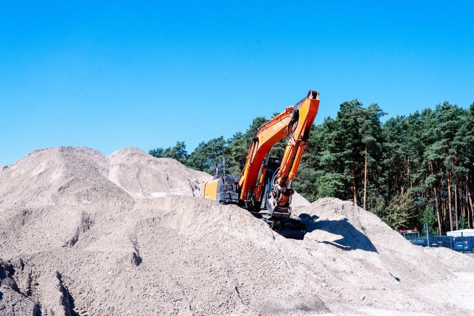 Orangefarbener Bagger auf einem Sandhaufen unter einem klaren blauen Himmel mit Kiefern im Hintergrund.