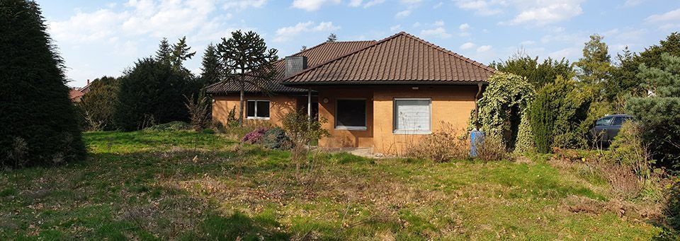 Ein kleines, orangefarbenes Haus mit einem braunen Ziegeldach steht in einem verwilderten Garten. Der Himmel ist blau mit weißen Wolken.