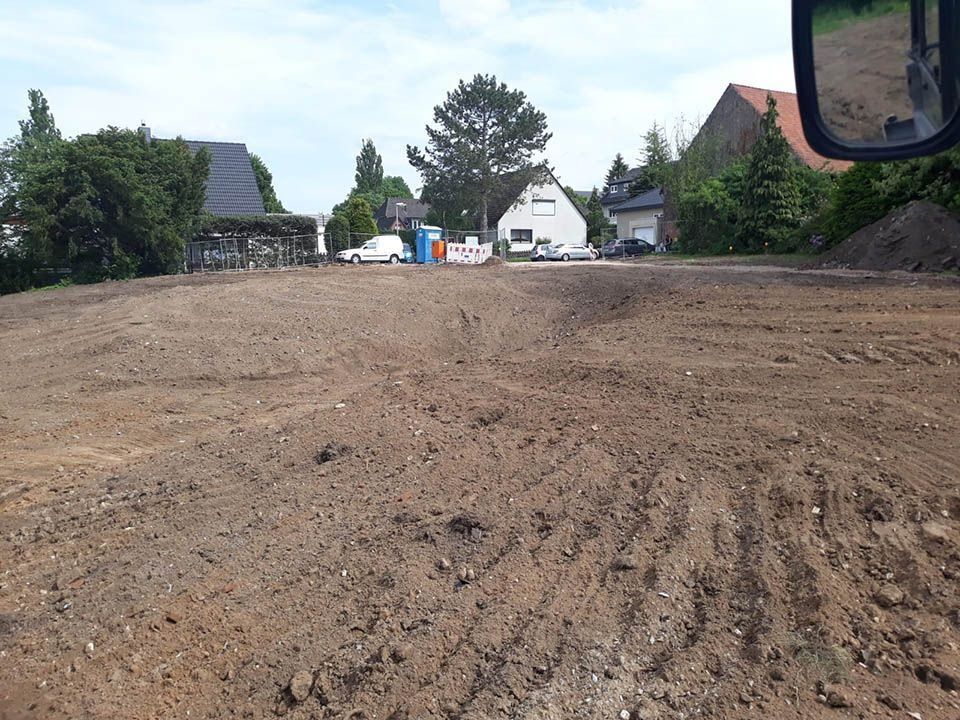 Geräumtes unbefestigtes Grundstück mit Baumaschinen im Vordergrund und Häusern im Hintergrund unter blauem Himmel.