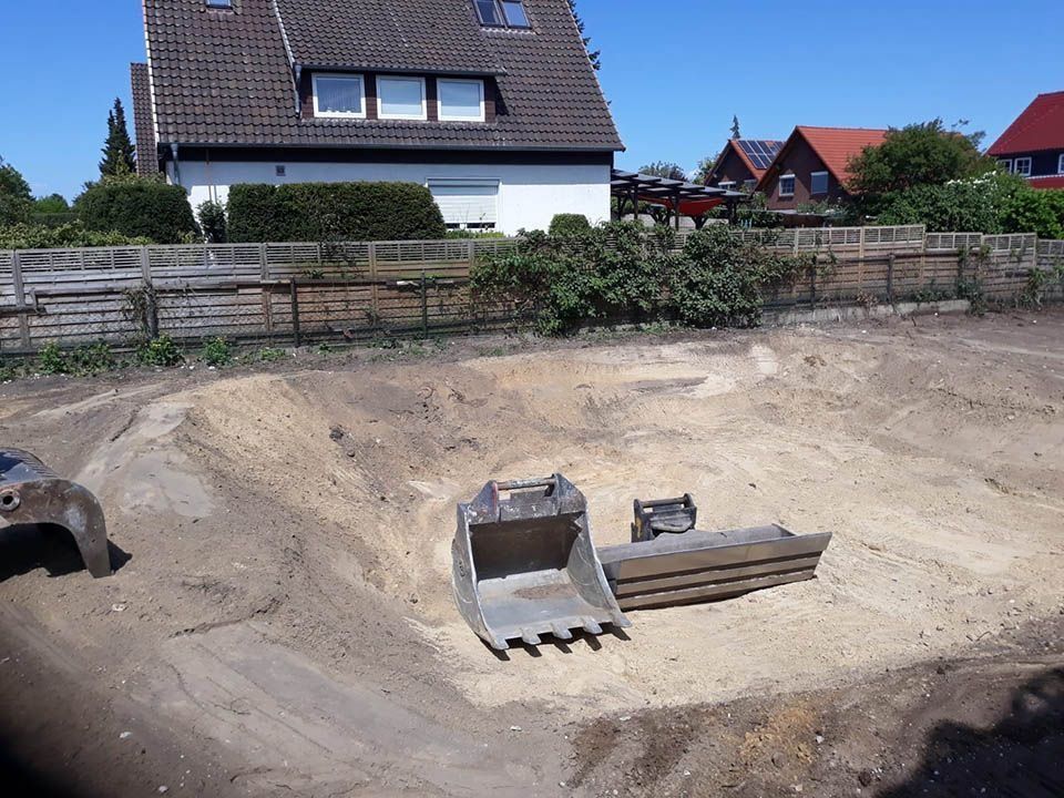 Bagger auf einer Baustelle, der Sand schaufelt, im Hintergrund sind Häuser zu sehen.