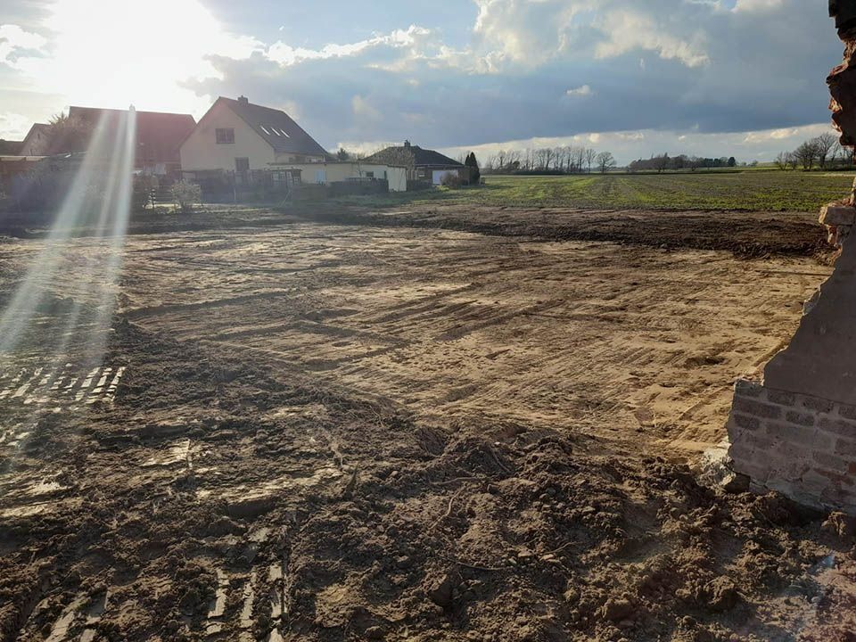 Schlammiges Feld mit entfernten Häusern unter bewölktem Himmel, im Vordergrund Reste einer Ziegelmauer.