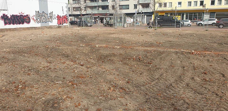 Ein leeres Grundstück mit Erde und einigen Baugeräten mit Gebäuden im Hintergrund.