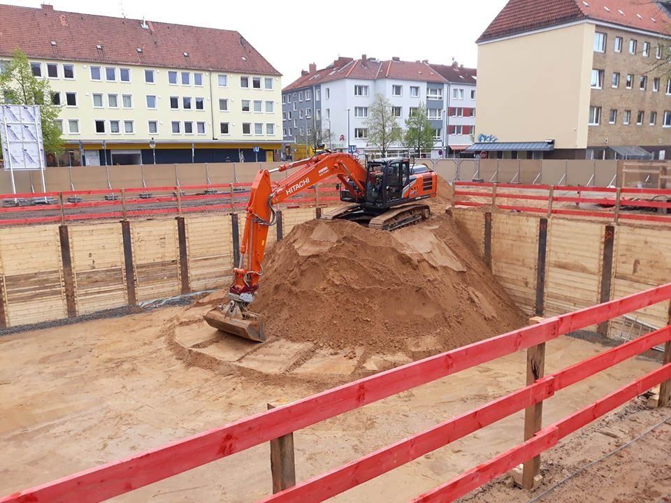 Baustelle mit einem orangefarbenen Bagger auf einem Sandhaufen, umgeben von Holzbarrieren und roten Zäunen, vor Wohnhäusern.
