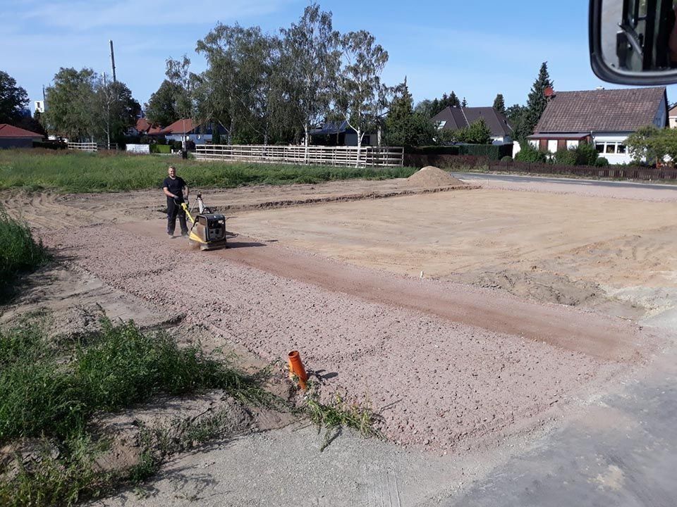 Mann verdichtet Kies auf einer Baustelle. Roter Kies auf einer geräumten Fläche, dahinter grünes Feld. Im Hintergrund Häuser.