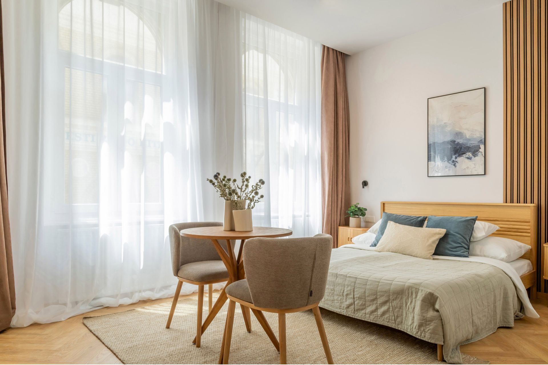 Cozy bedroom with sheer curtains, round table with chairs, bed, and artwork at Sora Suites Budapest.