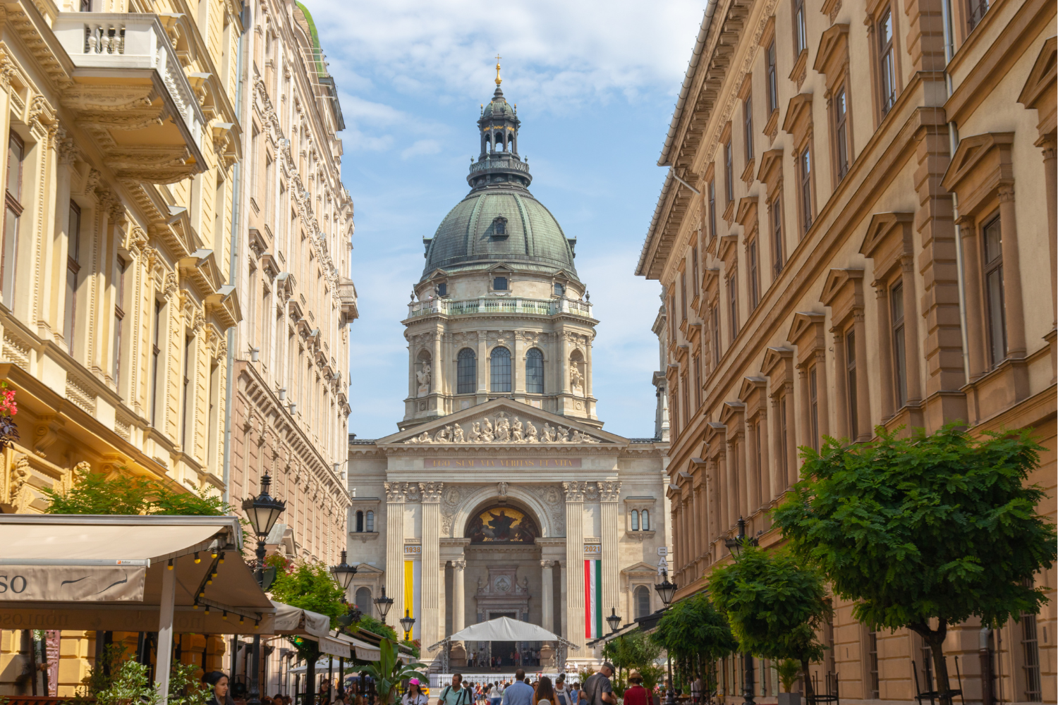 St. Stephen's Basilica, Budapest, viewed between buildings. Green dome, light-colored architecture, people, blue sky.