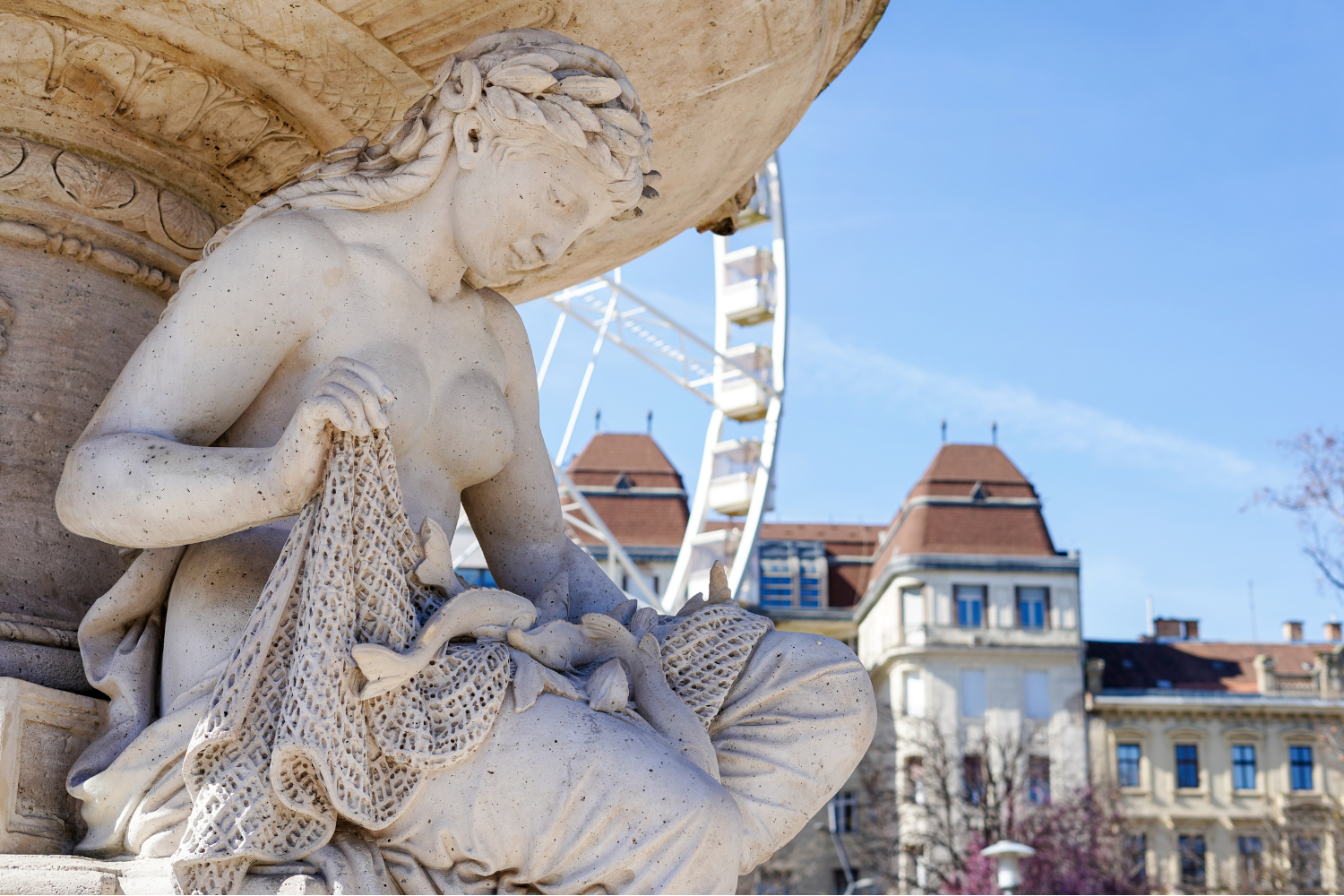 Statue of a woman seated, draped with netting, with the Budapest Eye Ferris wheel and building in the background. Sunny day.