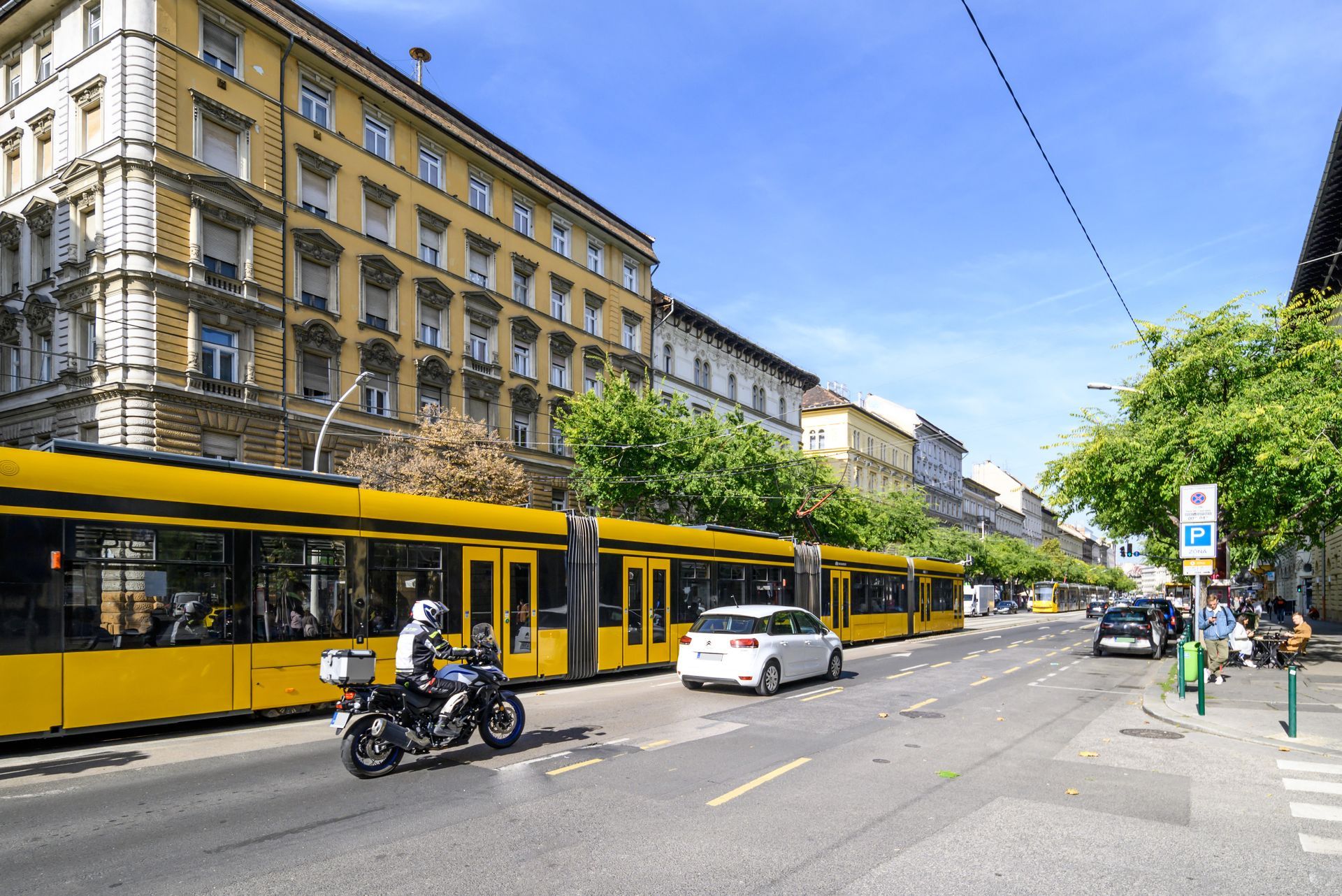 Yellow trams and traffic on the Great Boulevard of Budapest with buildings and trees under a sunny sky.