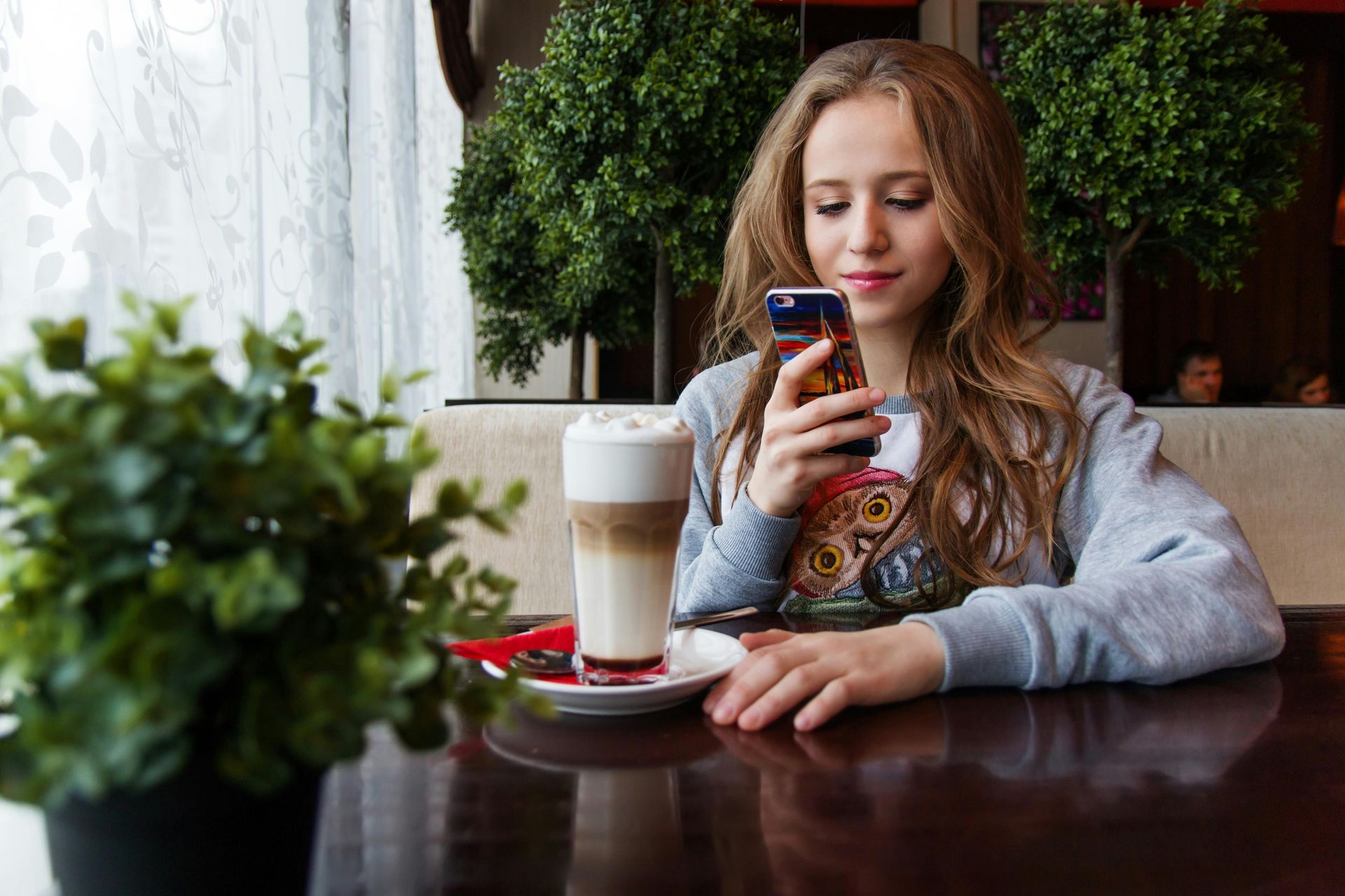Young woman in a cafe looking at her phone, next to a latte and plant.