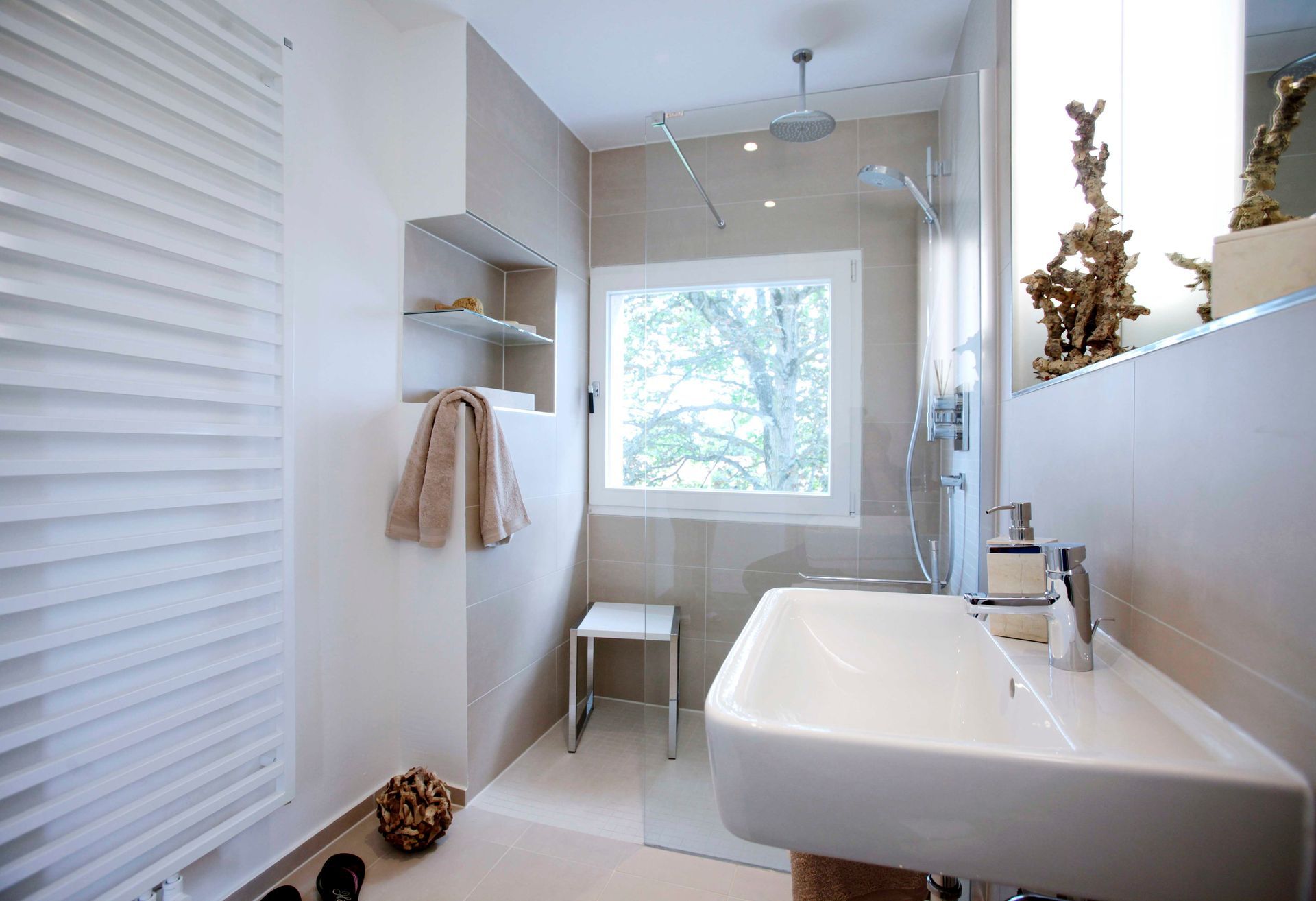 Ein helles, modernes Badezimmer mit weißem, wandmontiertem Waschbecken, verglaster Dusche und einem Hocker am Fenster.