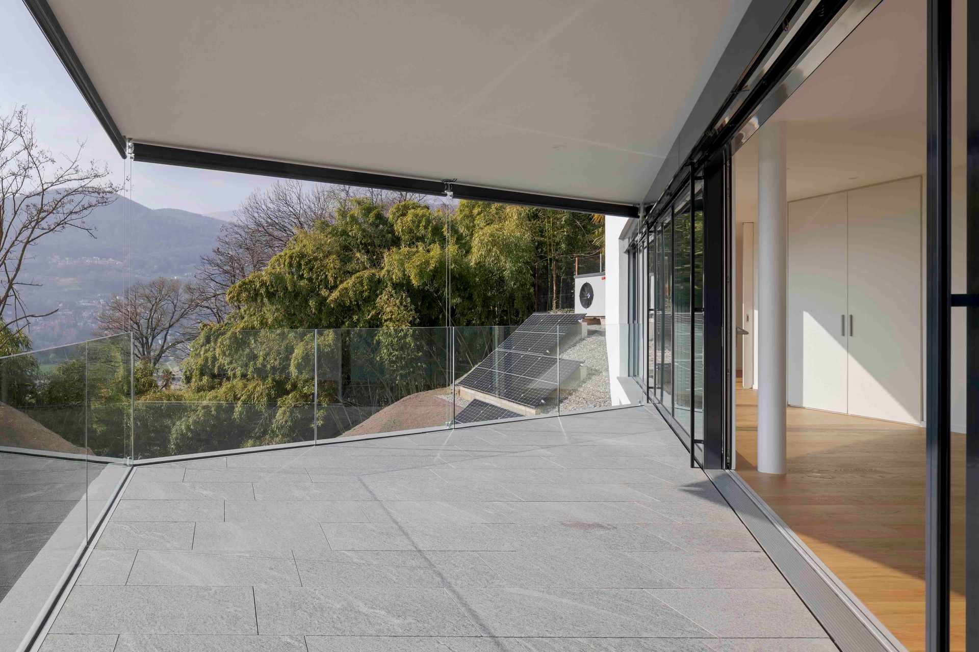 Eine Steinterrasse mit Glasgeländer bietet einen Blick auf die Berglandschaft und wird von einer Markise beschattet.