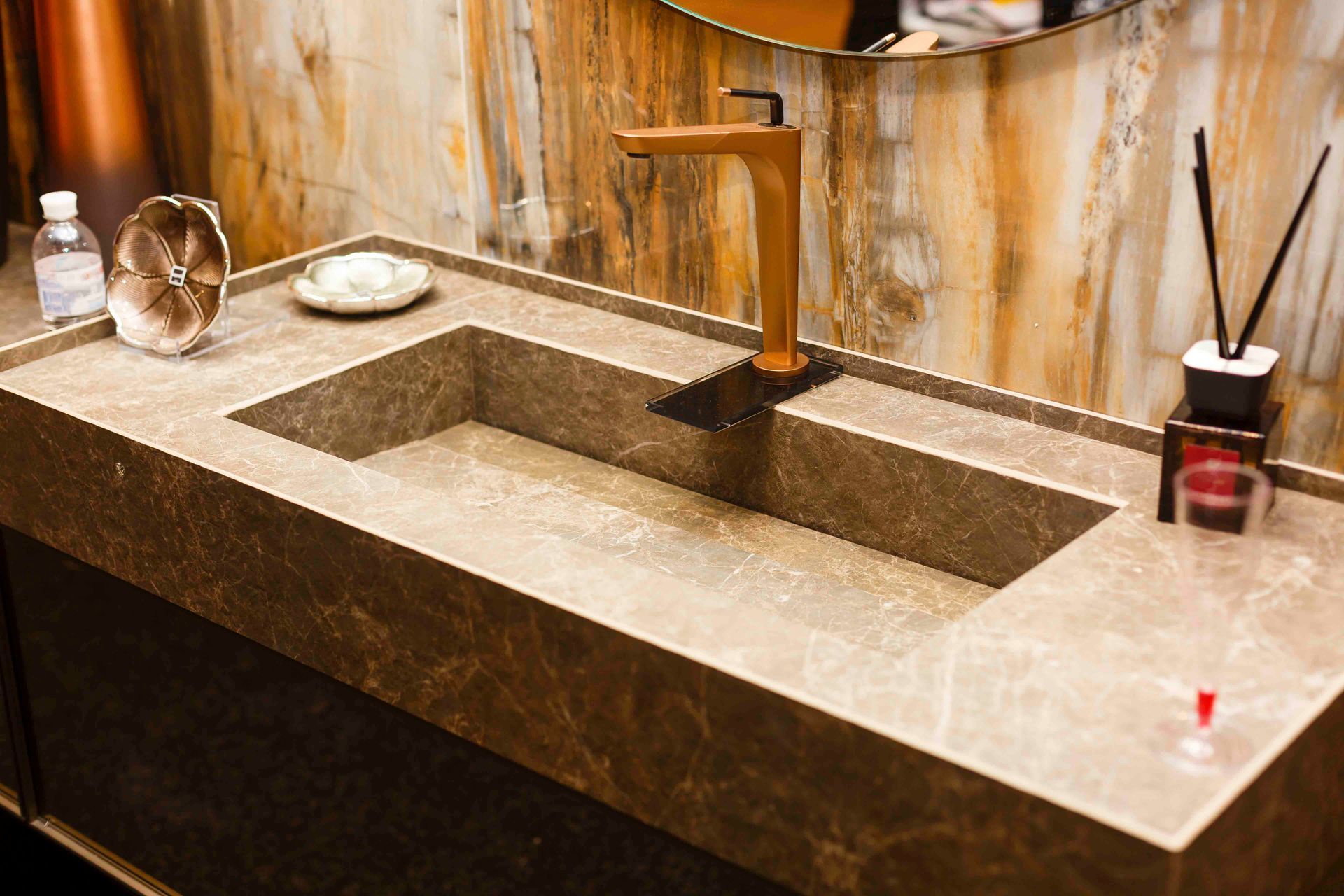 Ein moderner Badezimmerwaschtisch mit rechteckigem Steinwaschbecken und goldener Armatur vor einer Marmorwand.