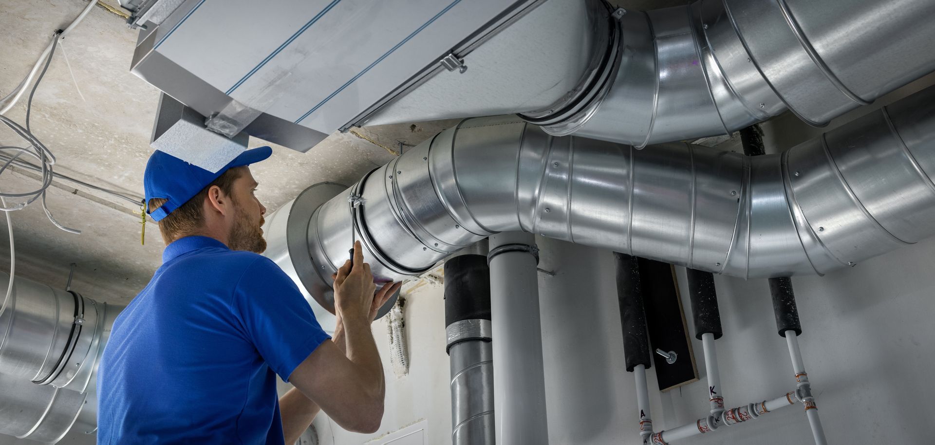 Un technicien CVC, vêtu d'une chemise et d'une casquette bleues, travaille sur des conduits d'air métalliques près du plafond.