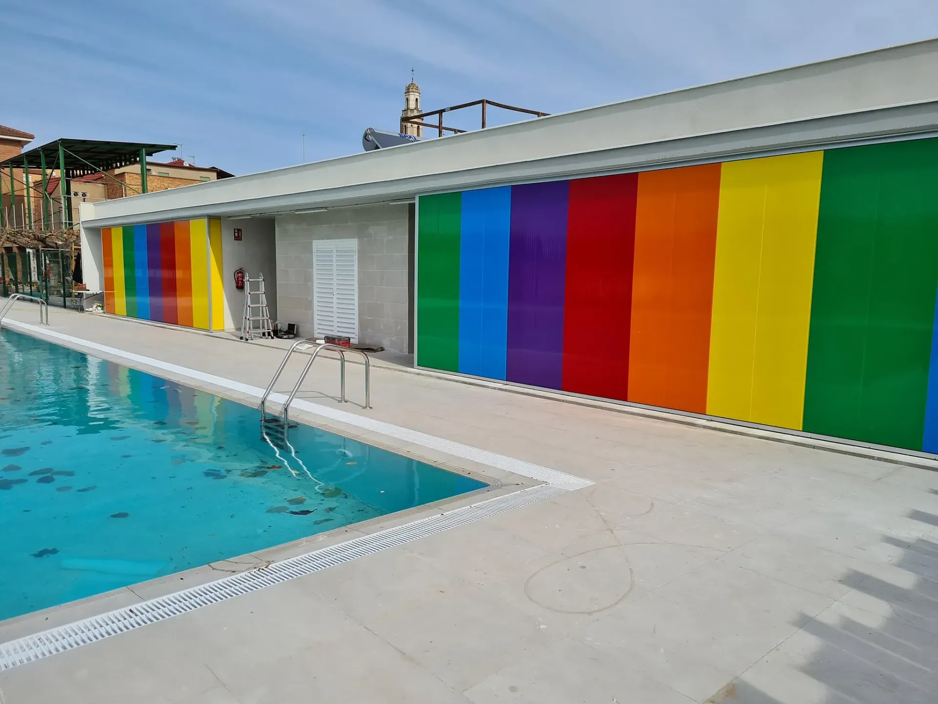 Una piscina rectangular con agua azul clara al lado de un edificio con paneles de colores con rayas de arcoíris.