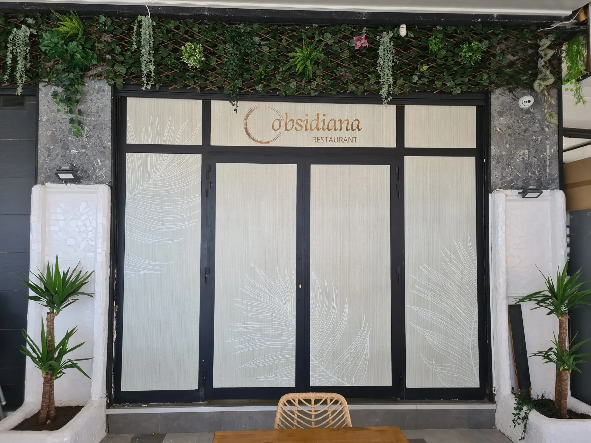 Entrada a un negocio llamado "Devasama". Cuenta con una puerta de vidrio con marco negro, rodeada de follaje verde y dos pequeñas plantas en maceta.