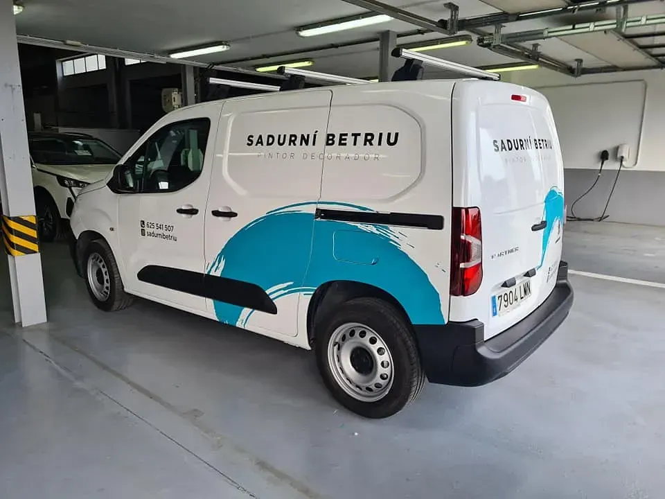Furgoneta blanca con diseño azul en un garaje. El texto "SADURNI BETRIU" aparece en el lateral y la parte trasera.