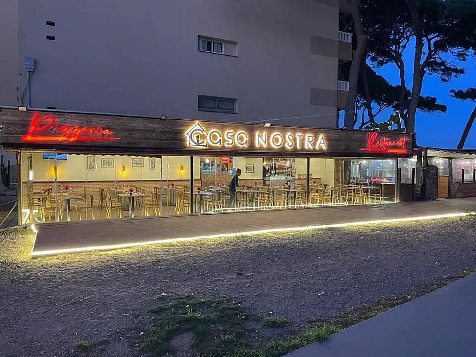 Restaurante Qeo Kosta al atardecer. Vista exterior de un edificio revestido de madera con letreros iluminados y mesas interiores visibles a través de las ventanas.