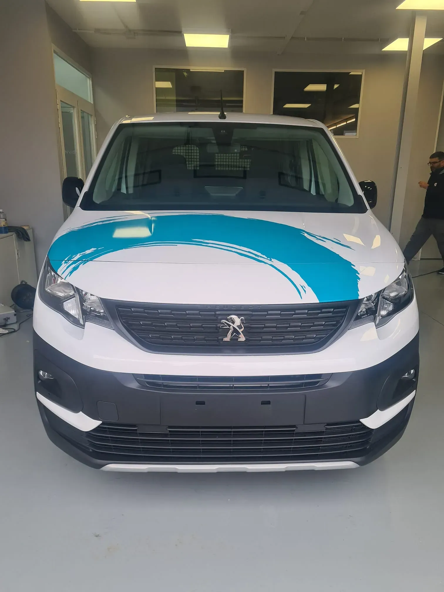Una furgoneta Peugeot blanca con un gráfico azul en el capó, estacionada en un garaje bien iluminado. Una persona camina al fondo.