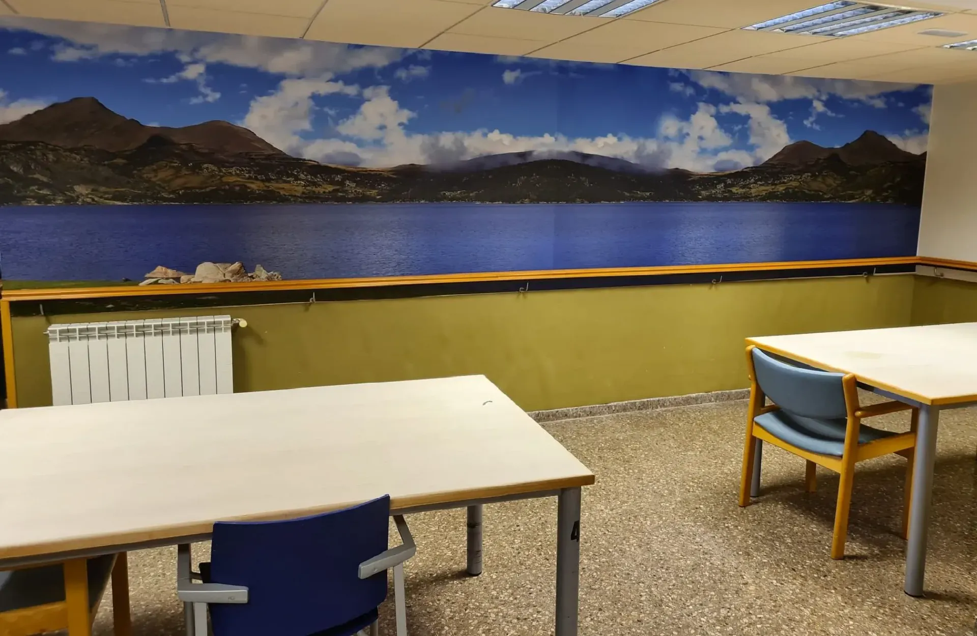 Una habitación con dos mesas y sillas frente a un mural que representa un lago y montañas bajo un cielo azul.