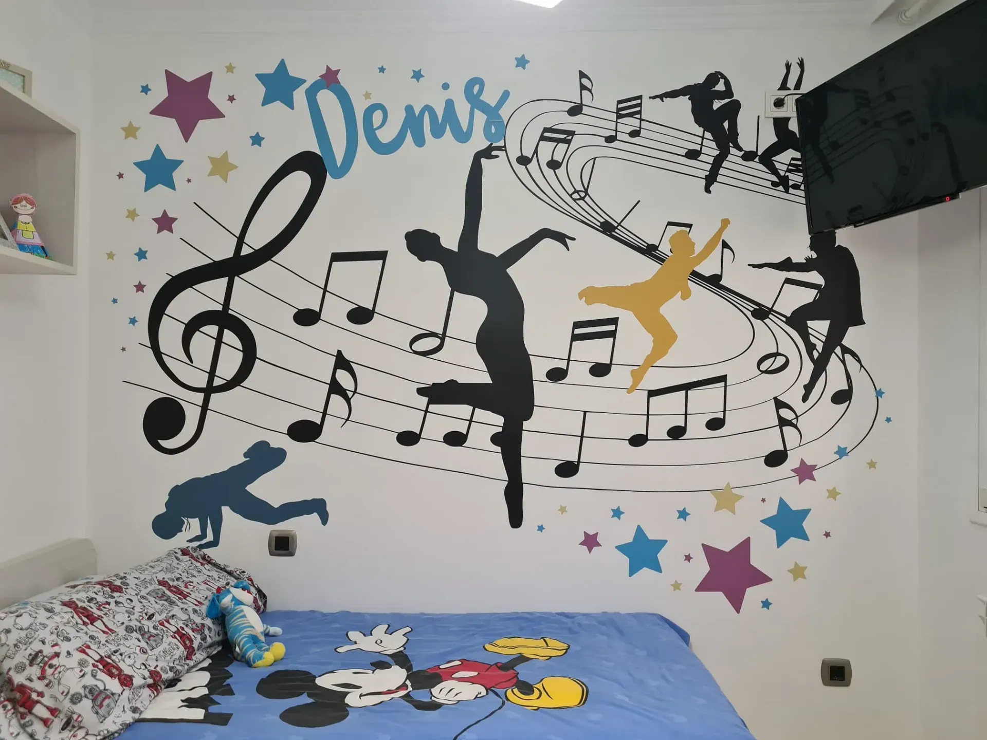 Una pared blanca con un mural de música y baile sobre una cama. El mural incluye notas musicales, estrellas y siluetas de bailarines.