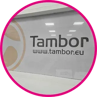 Logotipo de la empresa “Tambor” sobre una superficie de vidrio con la dirección del sitio web; borde circular rosa.