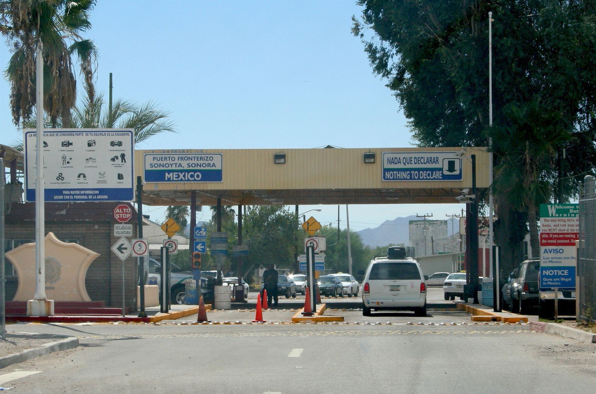Border checkpoint lane with cars, orange cones, and overhead signs on a sunny road