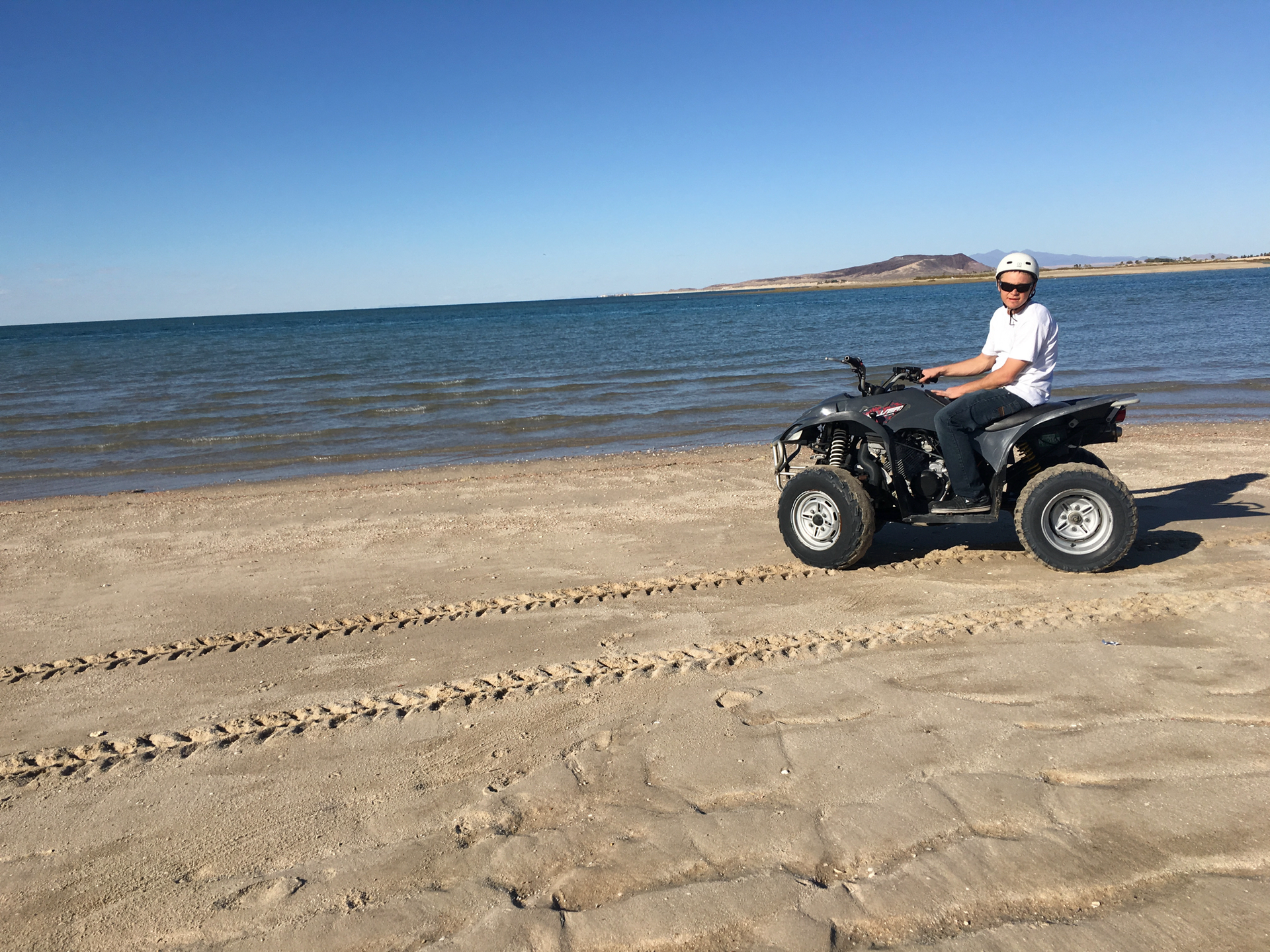 Person riding an ATV on a sandy beach beside the ocean under a clear blue sky