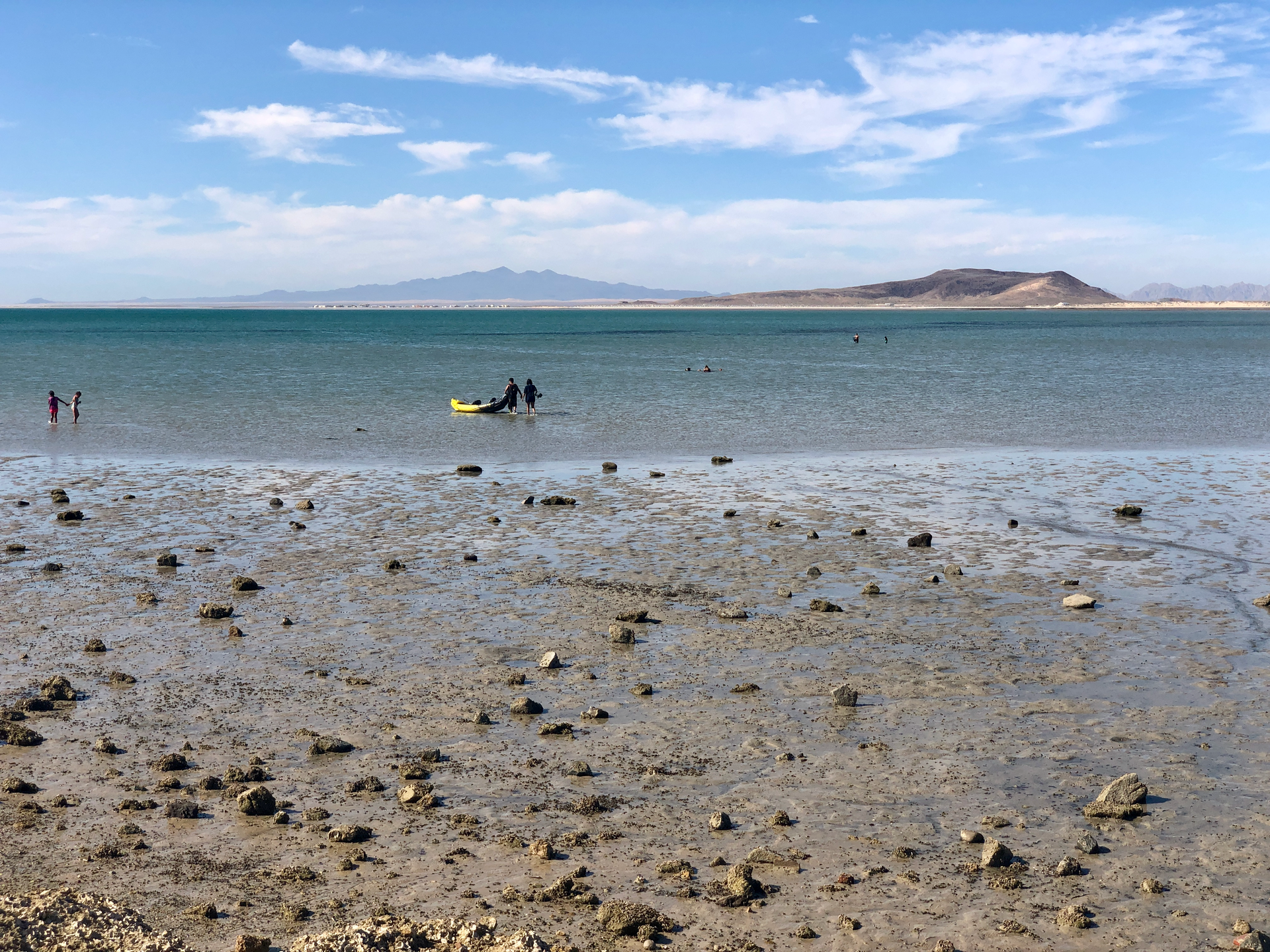 People wading on a rocky beach with a shallow blue sea and distant islands under a partly cloudy sky