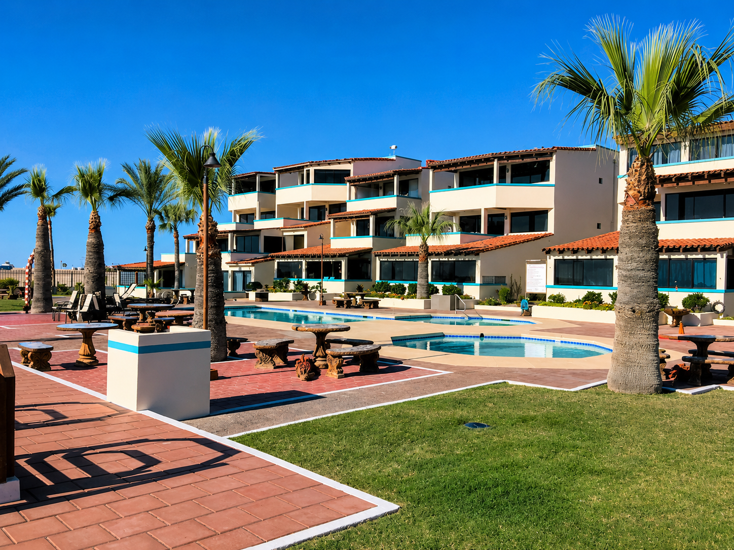 Sunny resort courtyard with palm trees, pools, lounge chairs, and a multi-story building under a blue sky