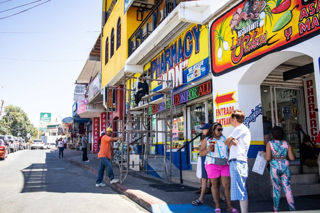 Colorful street storefronts with pedestrians on a sunny sidewalk beside parked cars
