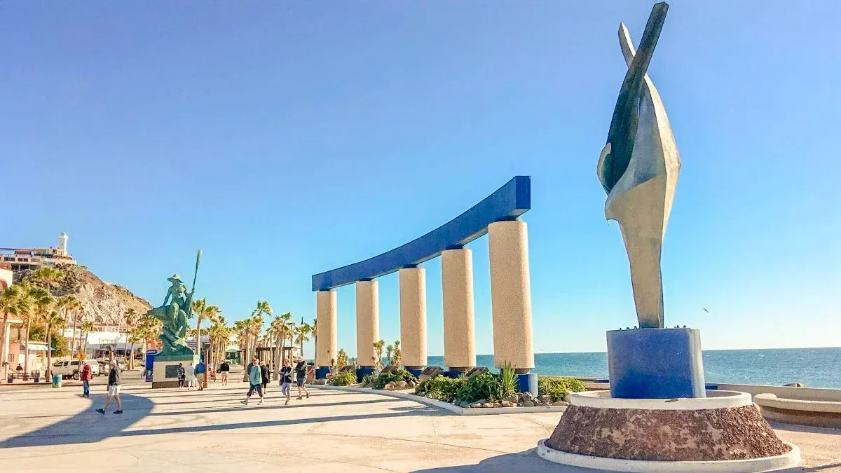 Modern seaside plaza with a blue arch sculpture, palm trees, and a bright sunny waterfront sky