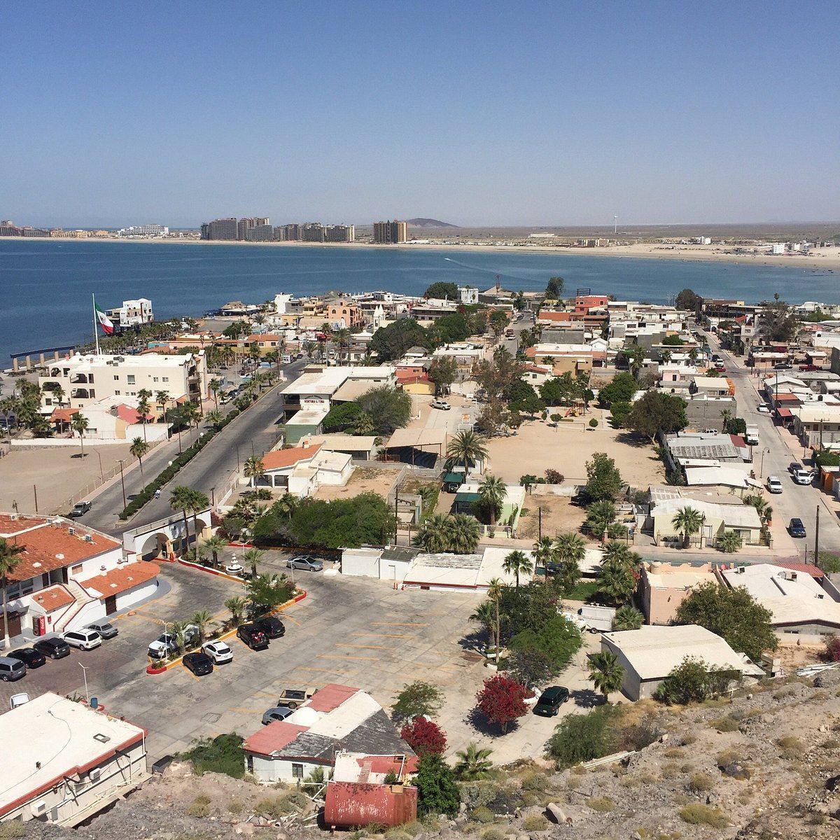 Coastal town with low buildings and roads beside a blue bay under a clear sky