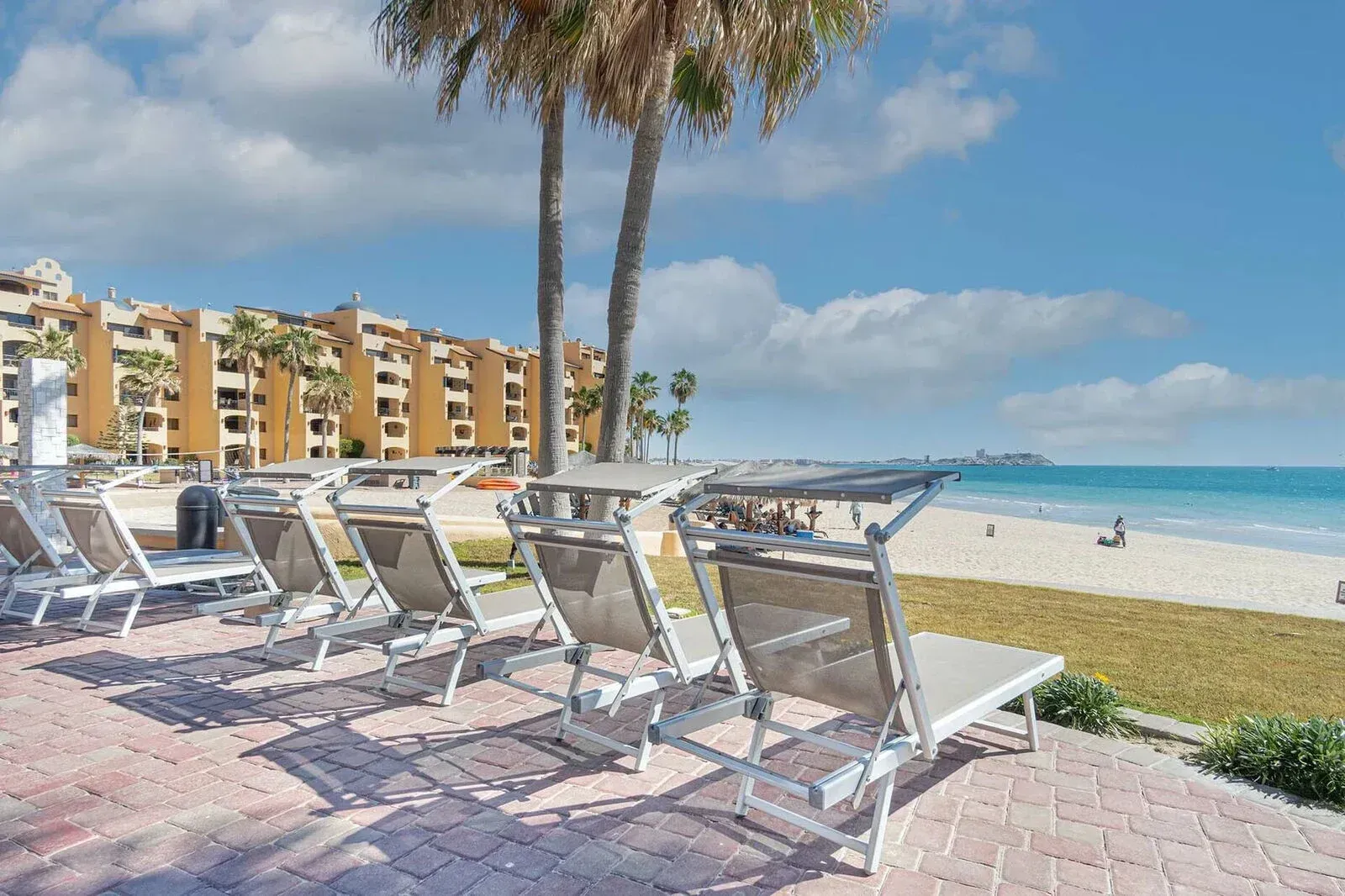 Beachfront lounge chairs under palm trees beside a sandy shore and blue ocean under a partly cloudy sky
