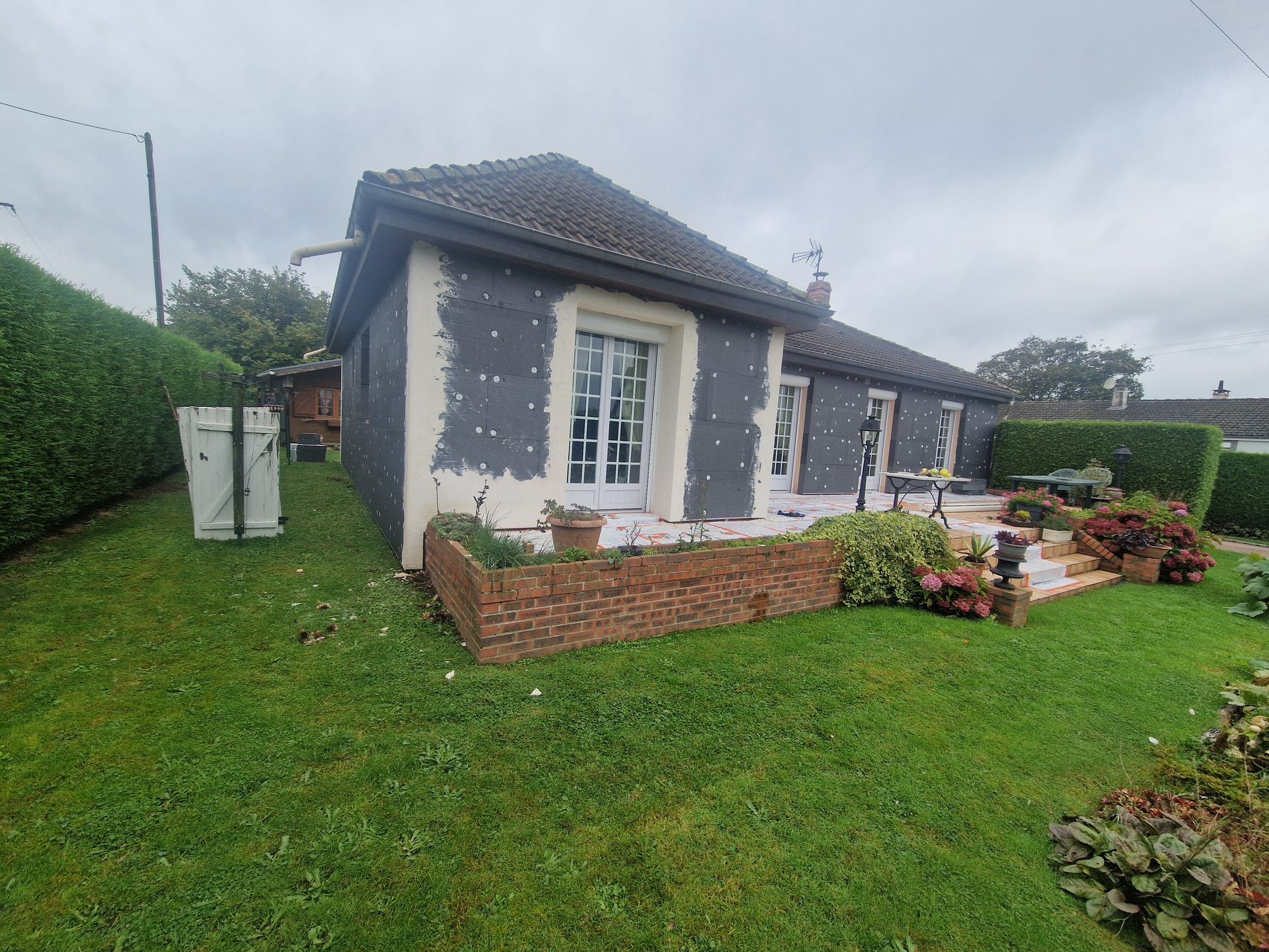 Maison aux murs gris en cours de rénovation extérieure, cour avant avec jardinière en briques et pelouse verte.