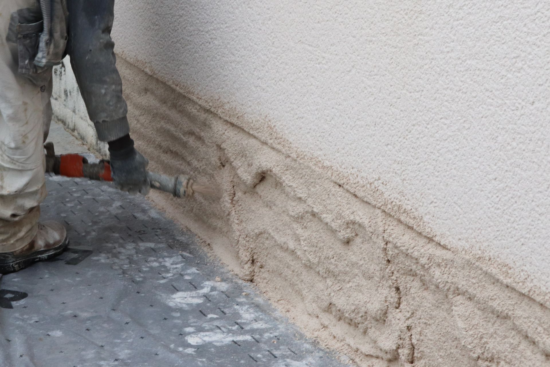 Une personne applique du stuc sur la base d'un bâtiment à l'aide d'un outil de pulvérisation ; vue extérieure.
