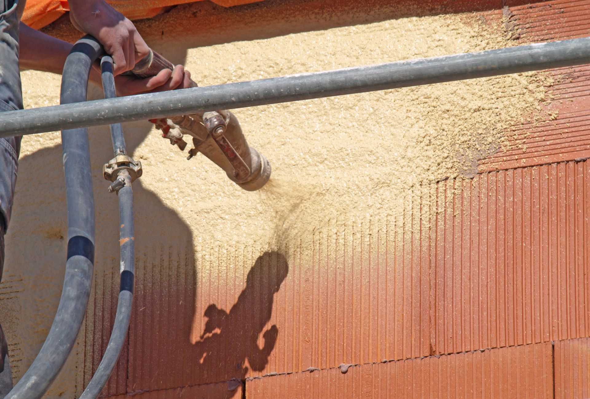 Un ouvrier pulvérise de l'isolant sur un mur de briques ; son ombre est visible.