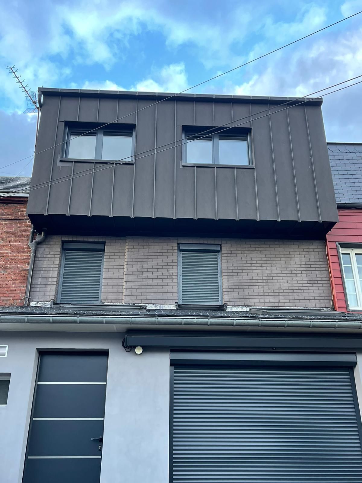 Bâtiment de deux étages à façade grise. L'étage supérieur comporte deux fenêtres. L'étage inférieur comporte deux fenêtres et une porte de garage.