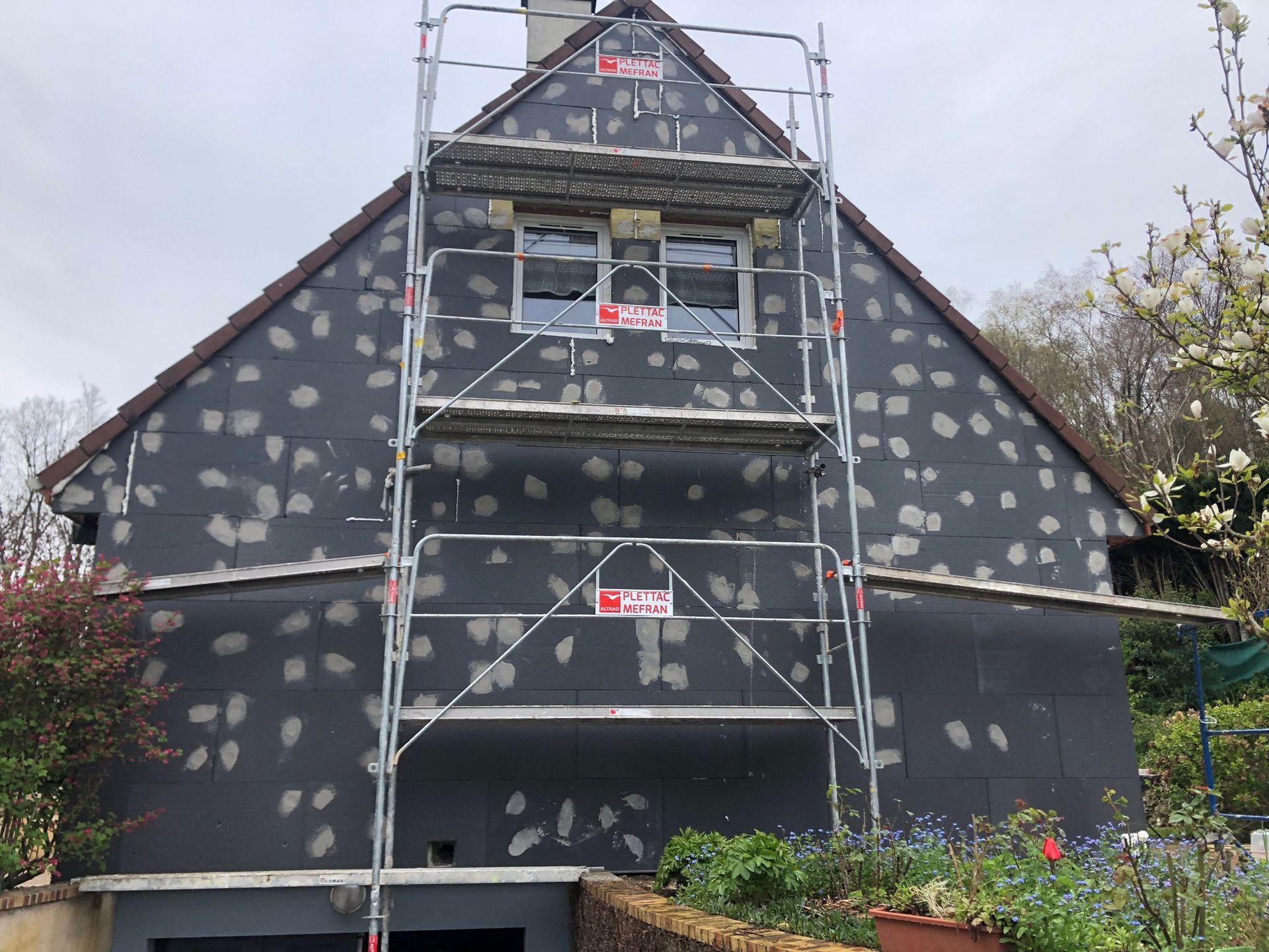Façade de la maison en rénovation ; échafaudages devant. Isolation noire tachetée de gris recouvrant le mur.