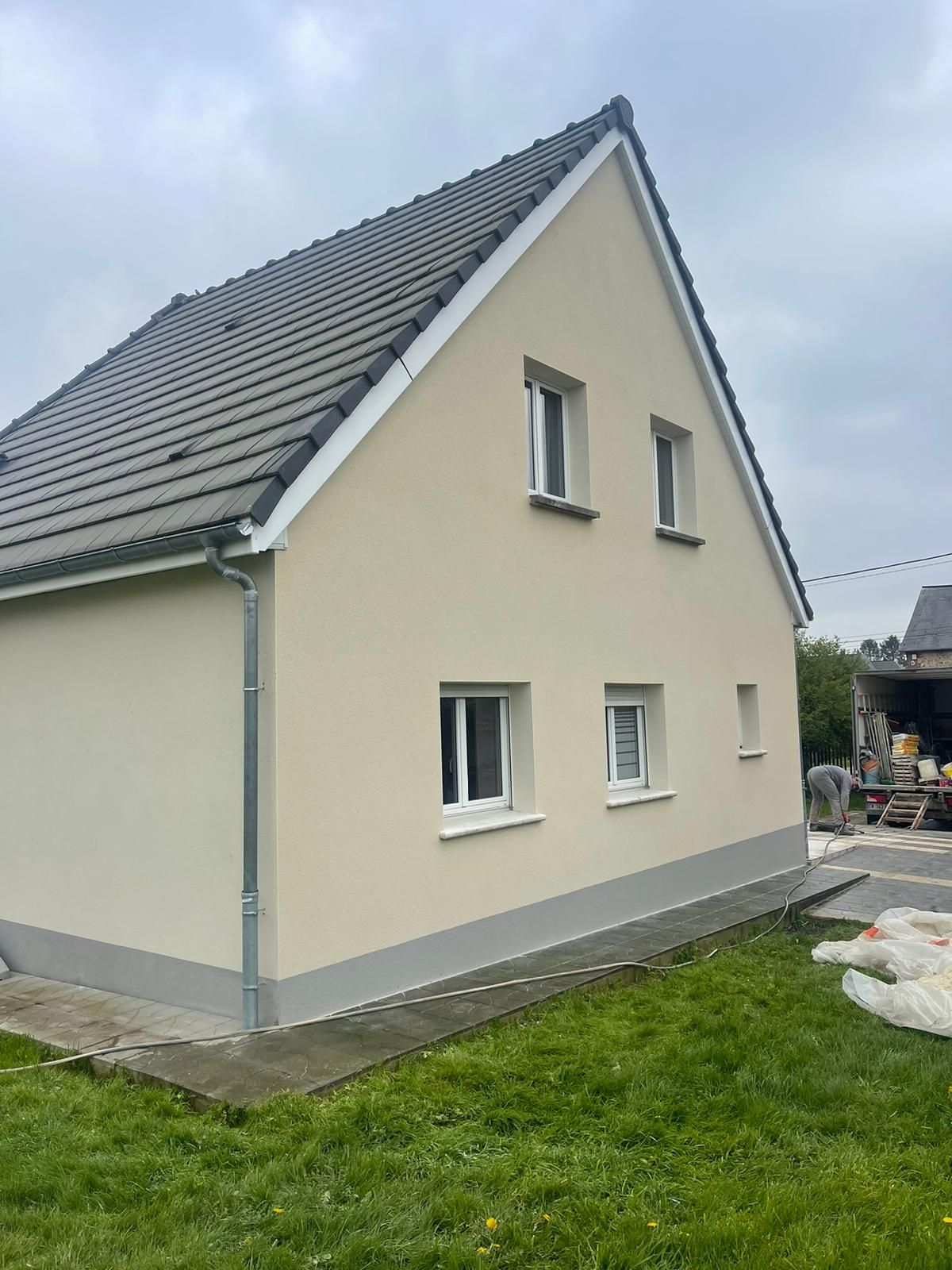 Maison beige avec toit et boiseries grises, encadrements de fenêtres blancs et pelouse verte.