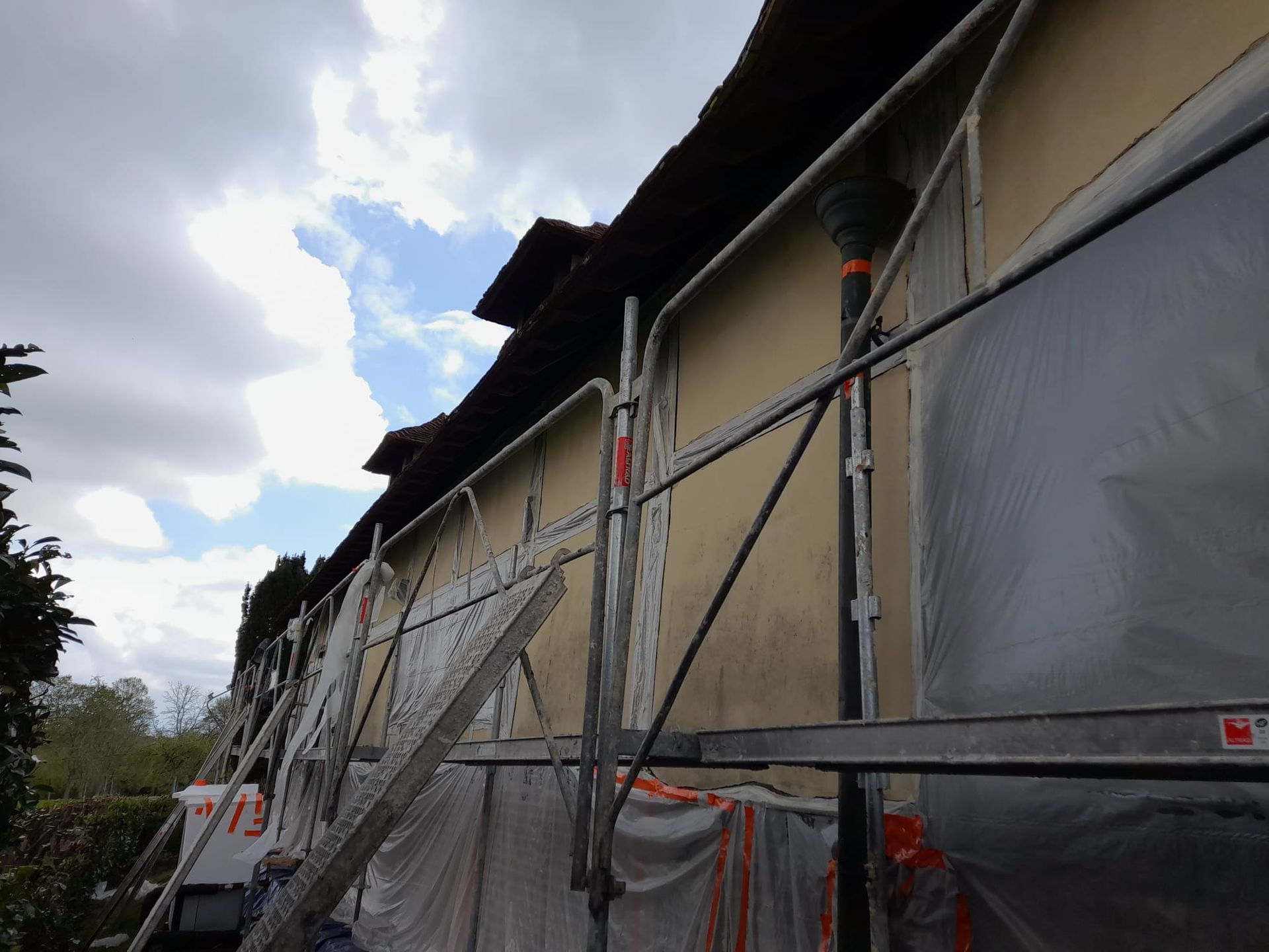 Échafaudages le long d'un bâtiment beige recouvert de bâches en plastique ; toit visible. Ciel nuageux au-dessus.