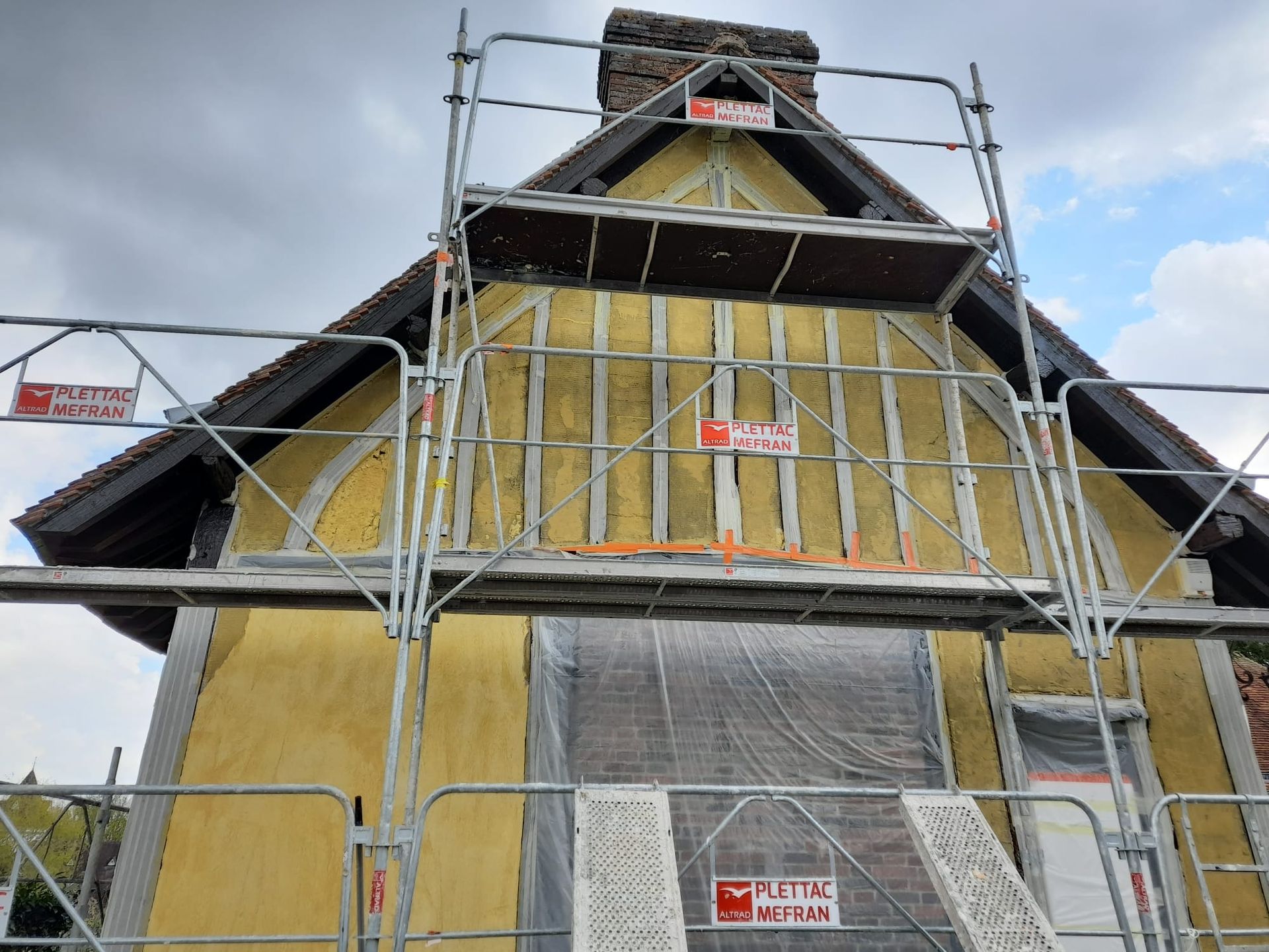 Façade de maison en rénovation, avec échafaudages. Extérieur jaune et marron, ciel couvert.