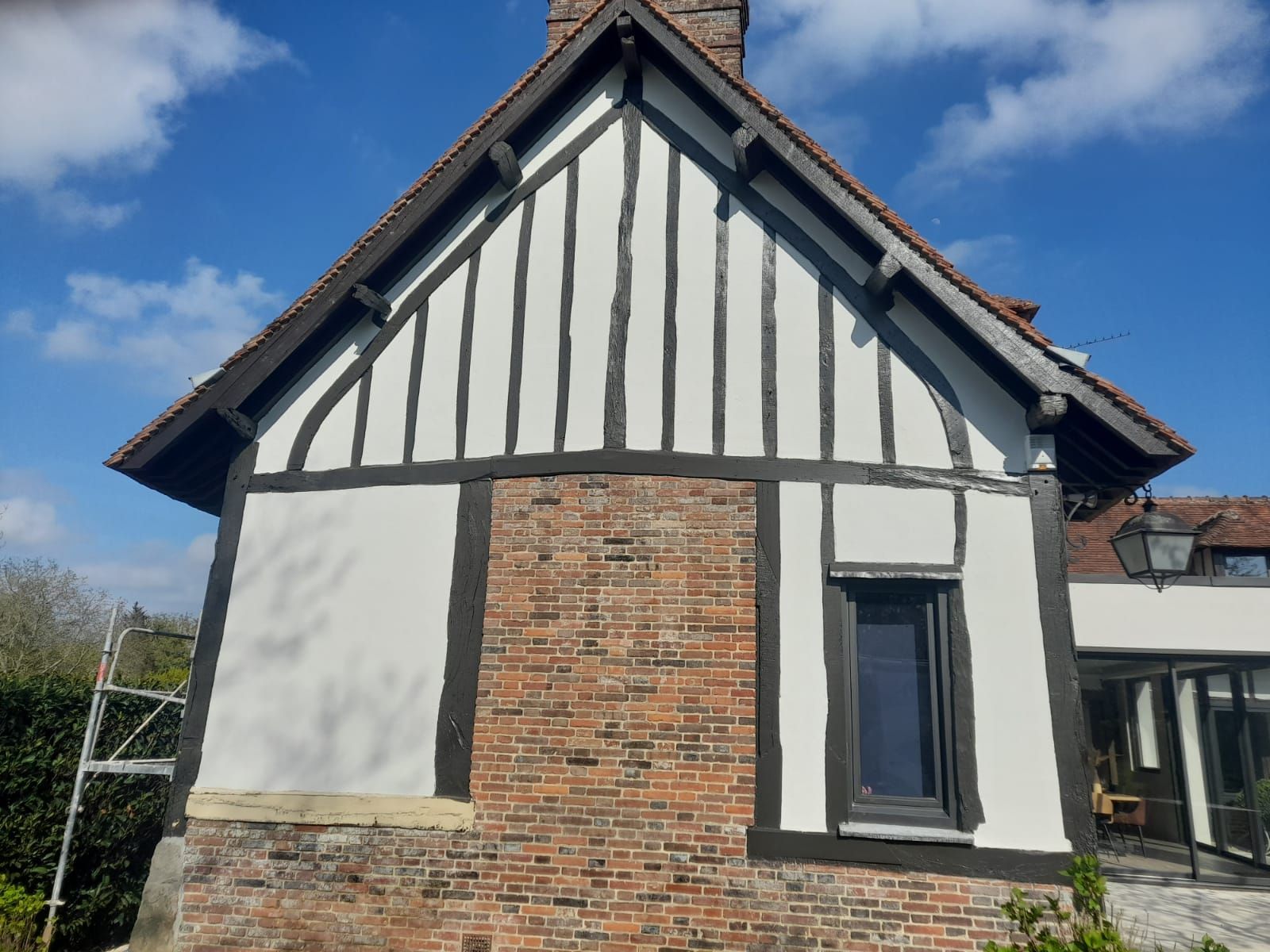 Vue latérale d'une maison aux murs enduits de plâtre blanc et aux poutres en bois sombre. On aperçoit la maçonnerie et une petite fenêtre.