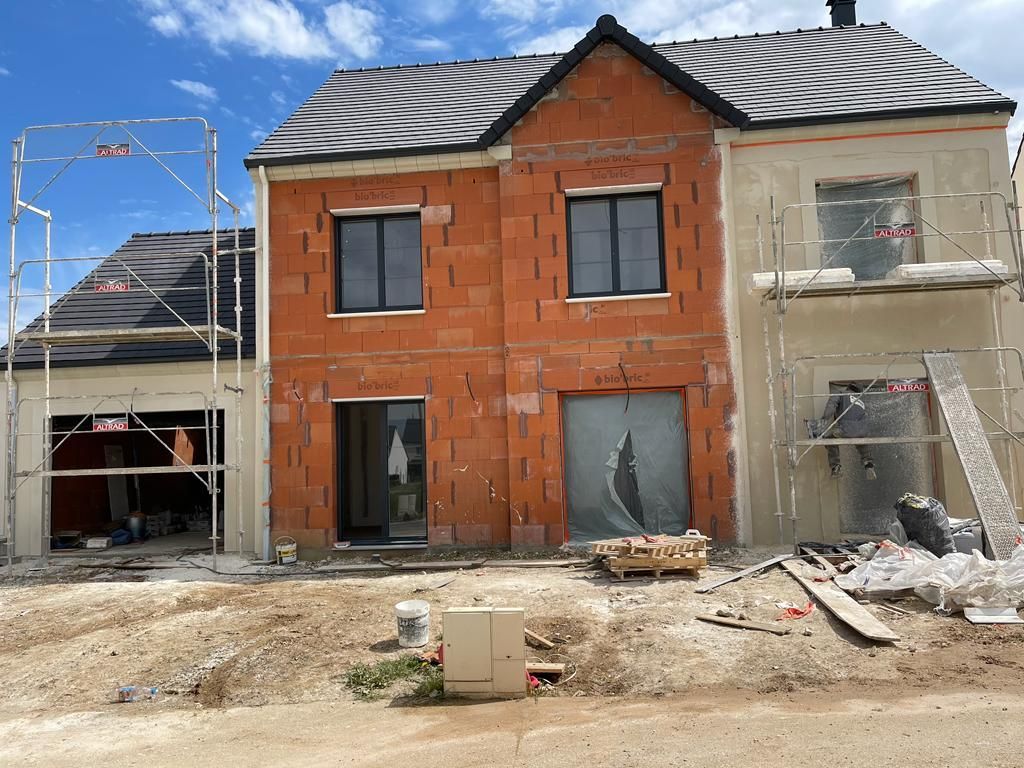 Maison à deux étages en construction, avec façade en briques apparentes et en stuc. Échafaudages visibles.