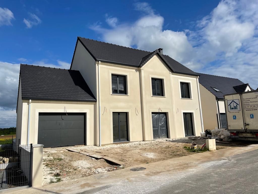 Maison beige neuve avec toit noir et garage. Aménagement paysager en cours ; ciel bleu ensoleillé.
